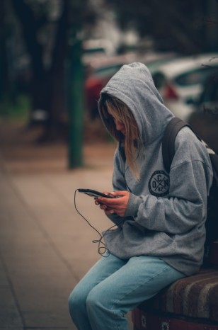 A teenager in a hooded sweatshirt, alone, looking down at a phone